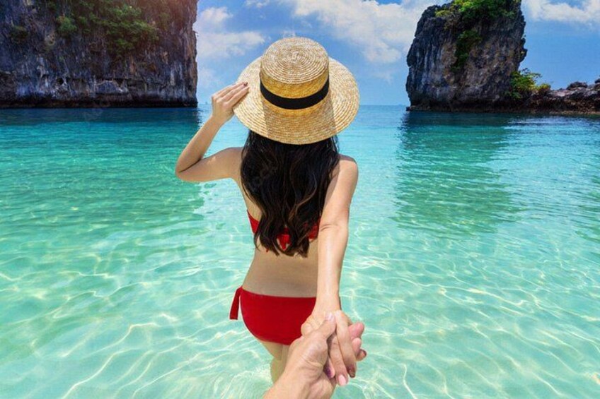 A person in a red bikini and straw hat holding hands with another person in clear turquoise water, with rocky cliffs in the background. 1