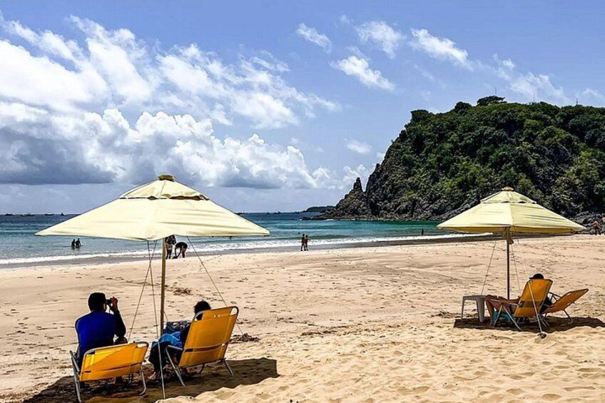 Una playa con dos sombrillas amarillas, gente relajándose y un acantilado rocoso al fondo. 3