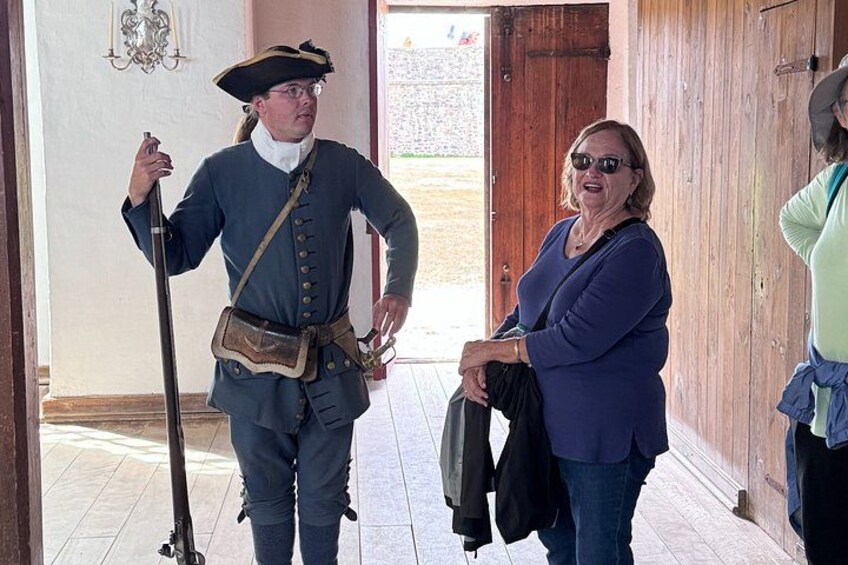 Eine Person in historischer Tracht mit einem Gewehr, eine andere Person hält eine Jacke, und im Hintergrund eine Holztür. 5