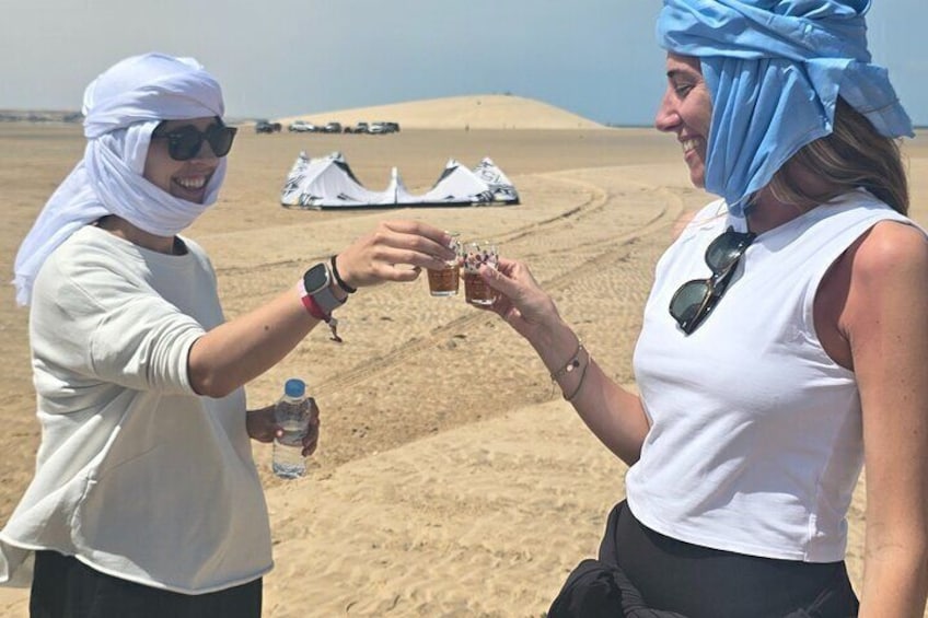 Two people toasting with drinks in a desert landscape with tents in the background. 1