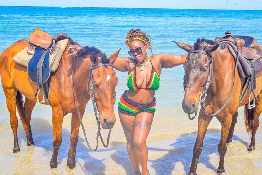 Eine Person in einem Bikini steht zwischen zwei Pferden an einem Strand, im Hintergrund das Meer. 4