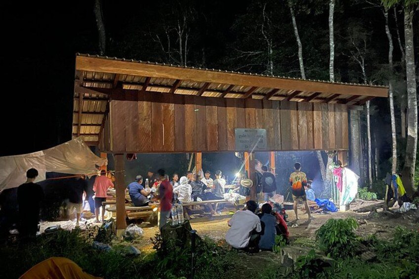 An outdoor gathering with people under a wooden shelter, surrounded by trees. 2