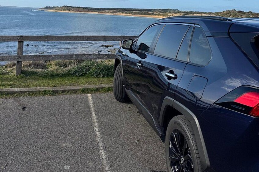 Un SUV azul oscuro estacionado en una carretera pavimentada con una cerca de madera y una playa al fondo. 2