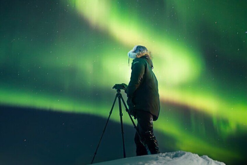A person with a camera on a tripod, standing on a snowy surface, looking at the Northern Lights. 3