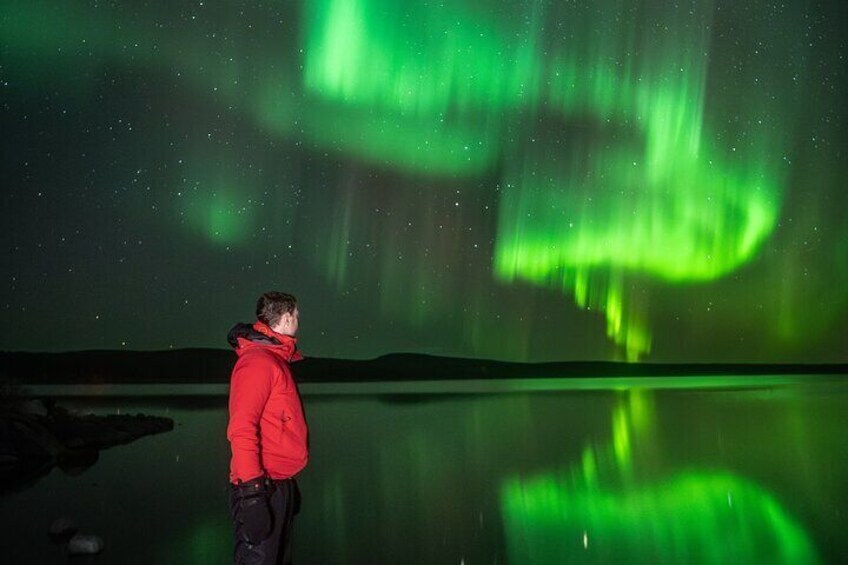 A person in a red jacket standing by a calm body of water, gazing at the night sky where the aurora borealis is visible. 2
