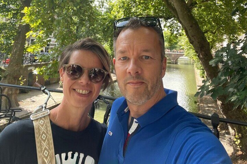 Twee personen maken een selfie langs een gracht met bomen en een brug op de achtergrond. 3