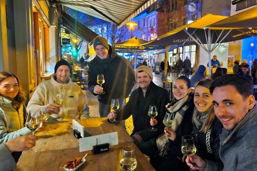 Un grupo de personas brindando con copas de vino en una mesa de madera, en un ambiente al aire libre con un corredor cubierto y letreros iluminados al fondo. 3