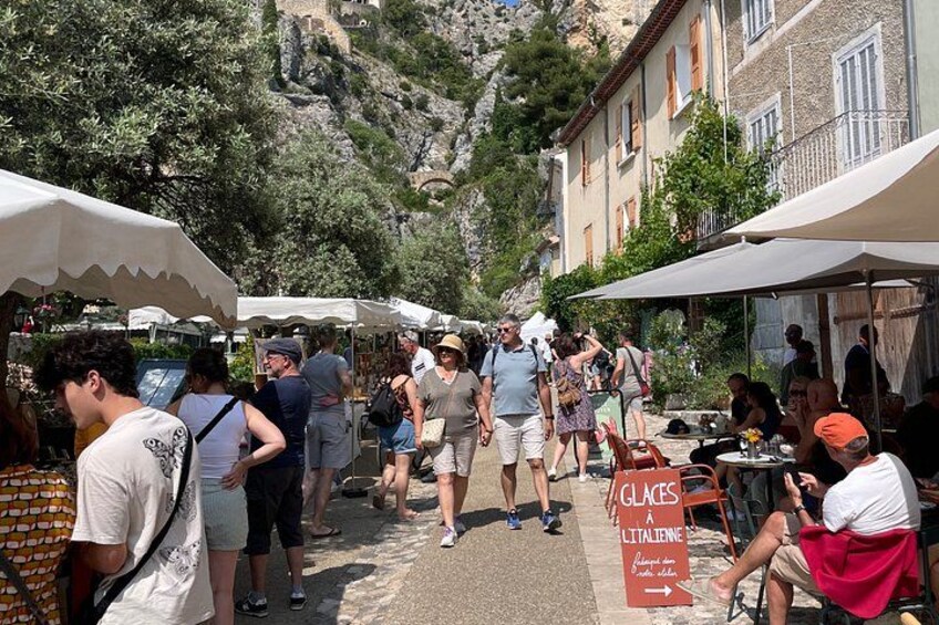 A crowded outdoor market with people walking and sitting under umbrellas, with buildings and trees in the background. 4