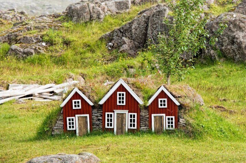 Trois petites maisons rouges aux contours blancs, situées dans un champ verdoyant parsemé de rochers et agrémenté d’un arbre. 3