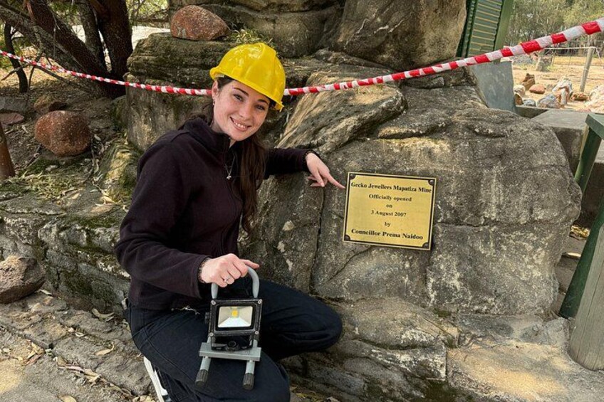Una persona con casco amarillo sostiene un dispositivo, sentada junto a una roca con una placa. 4