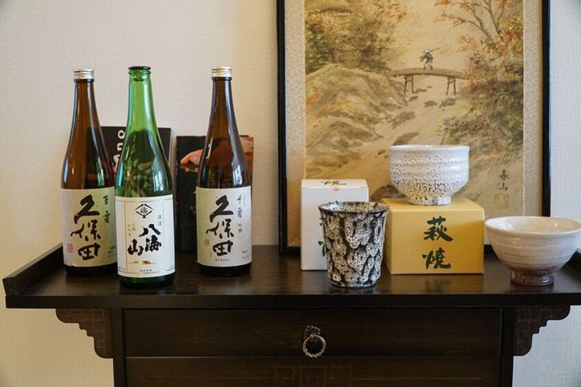 Three bottles of sake, a ceramic bowl, and a painting on a shelf. 4