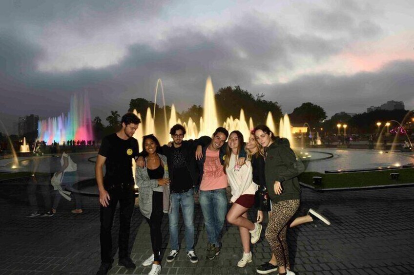 A group of people posing in front of a fountain with colorful lights at dusk. 1