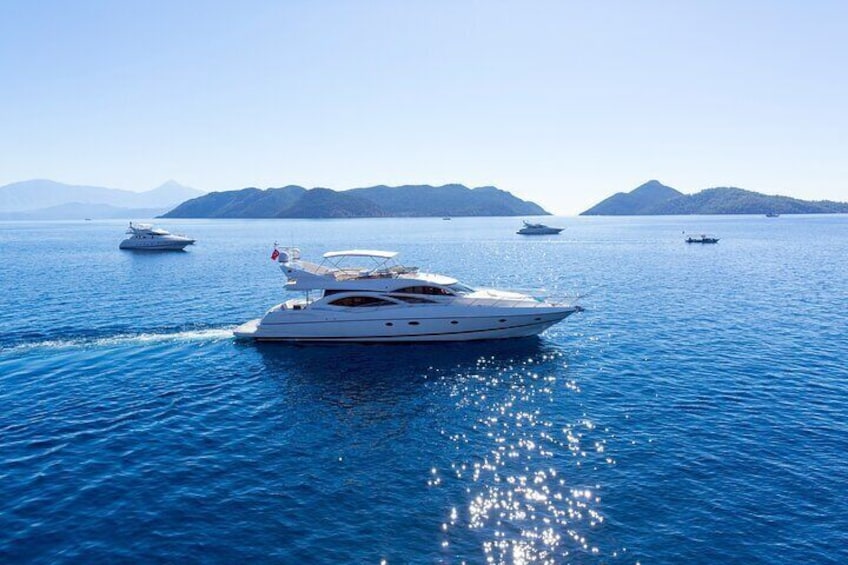A large yacht on calm blue waters with several smaller boats in the background and distant mountains. 1