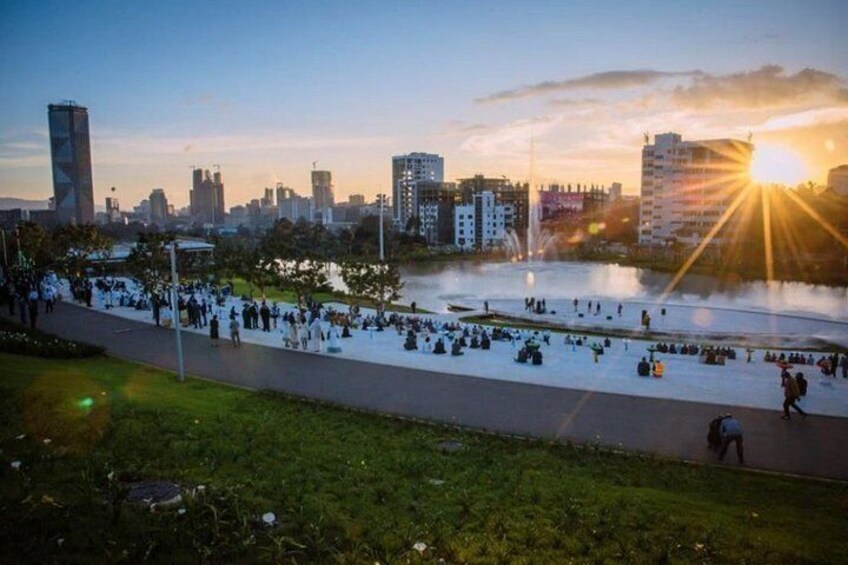 Un parque junto al río con gente caminando y sentada, un atardecer y un horizonte urbano al fondo. 2