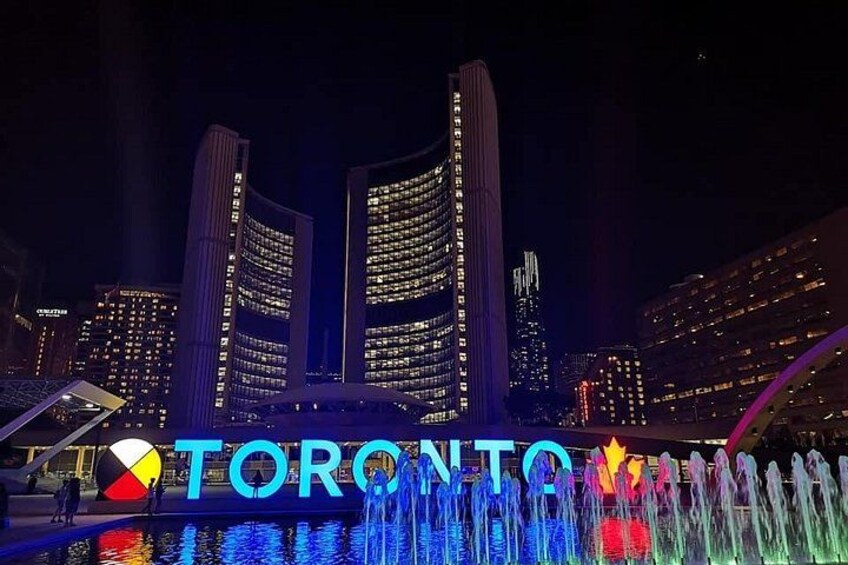 A nighttime cityscape with illuminated buildings and a water fountain display in front of a sign reading 'TORONTO'. 3