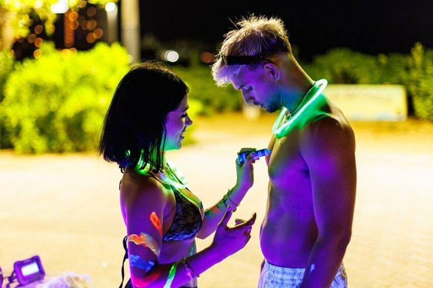 Two people, one with a neon light around their neck, standing in an outdoor area with greenery and a building in the background. 2