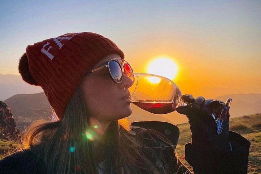A person wearing a red beanie and sunglasses, holding a glass of red liquid, with a sunset in the background. 4