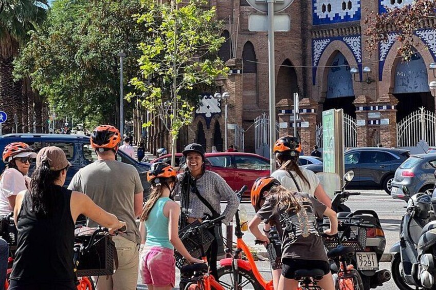 A group of people with bicycles and helmets, a red car, and a building with arches in the background. 2