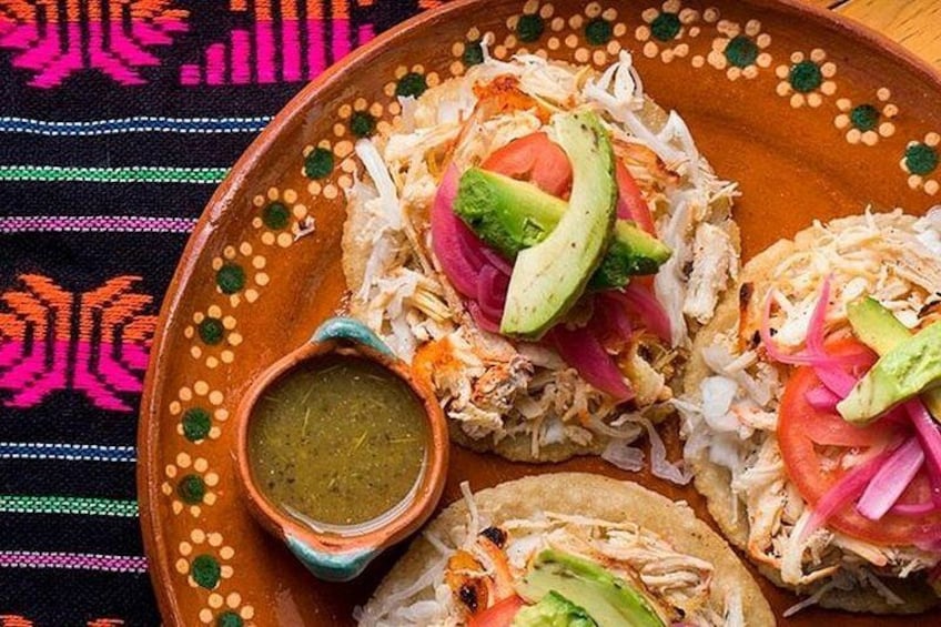 A plate of tacos with avocado, tomato, and onion toppings, and a small bowl of green sauce. 3