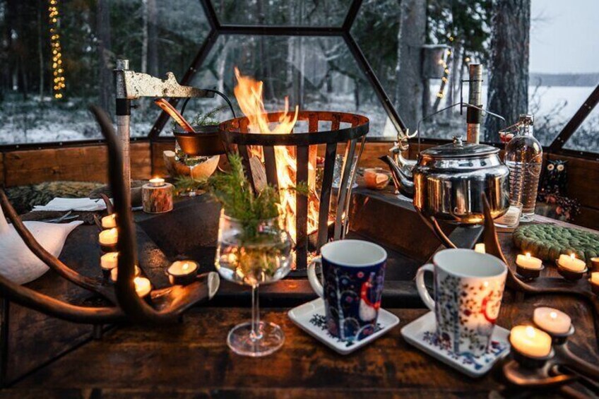 En mysig stuga med en vedeldad kamin, ljus och muggar på ett bord. 3