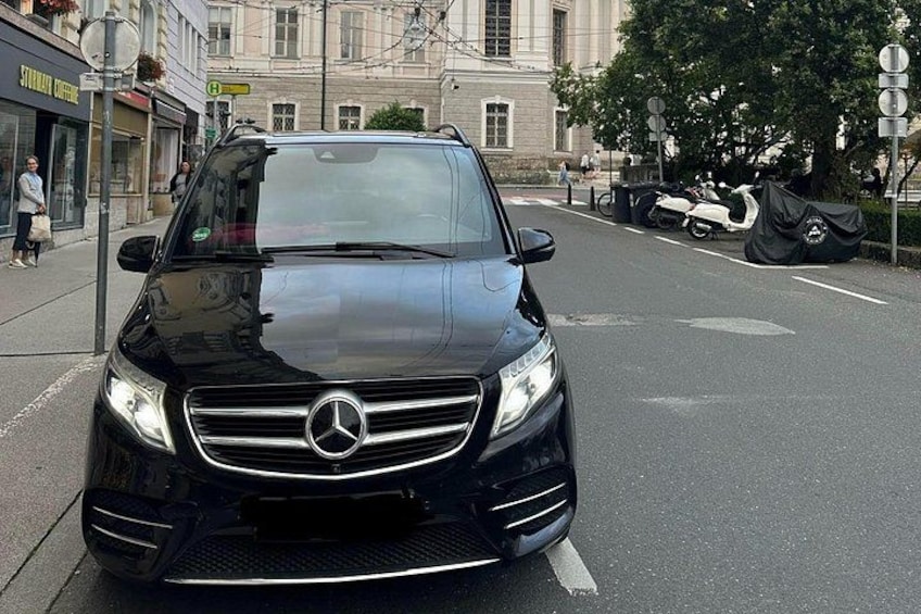 Een zwarte auto geparkeerd op een stadsstraat, met gebouwen en een boom op de achtergrond. 4