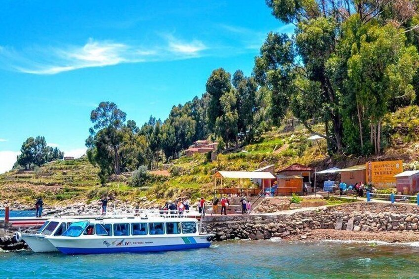 Un barco amarrado en la orilla, con gente en la costa, un muro de piedra y una colina con edificios al fondo. 5