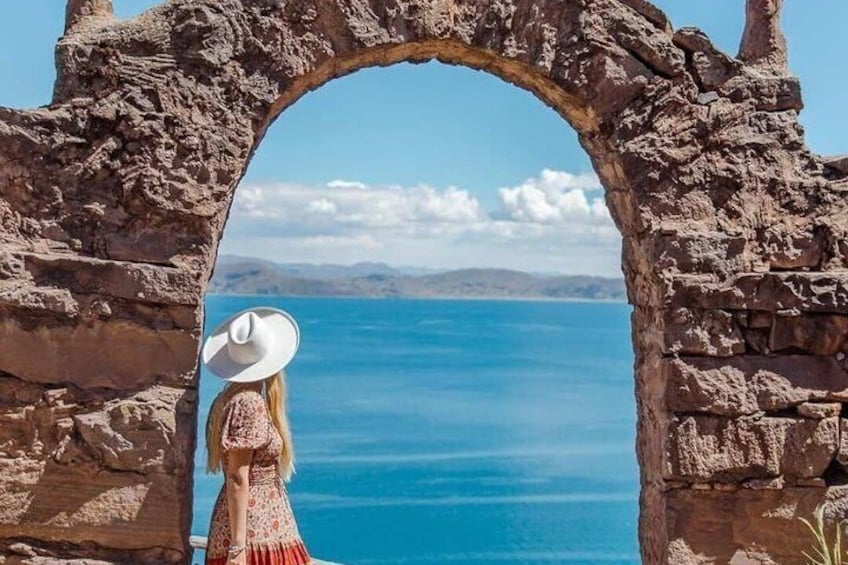Una mujer con sombrero blanco y vestido rojo posa frente a un arco de piedra con vistas a un cuerpo de agua. 1