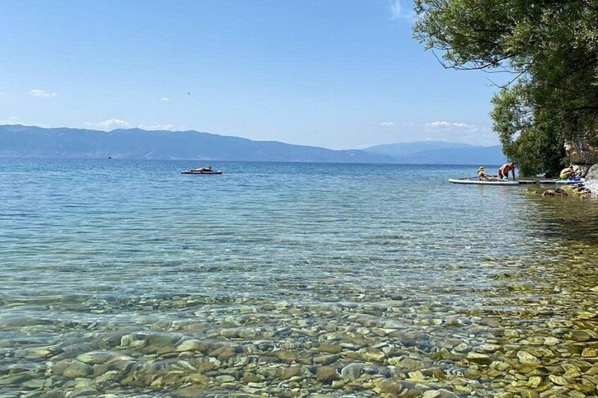 A clear lake with rocks at the shore, a boat in the distance, and people sitting on the rocks. 4