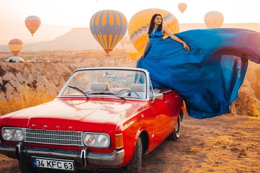 En rød, klassisk bil med nummerplade, en person i en flagrende blå kjole, og luftballoner i baggrunden. 5