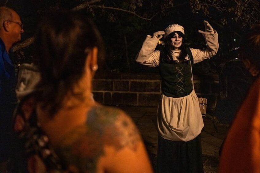Una persona vestida con traje de época, con las manos levantadas, posiblemente actuando o gesticulando, en un entorno exterior tenuemente iluminado. 5