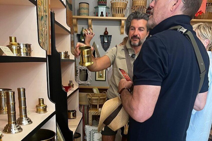 A person holding a brass container, another person in a beige shirt, and shelves with various items. 5