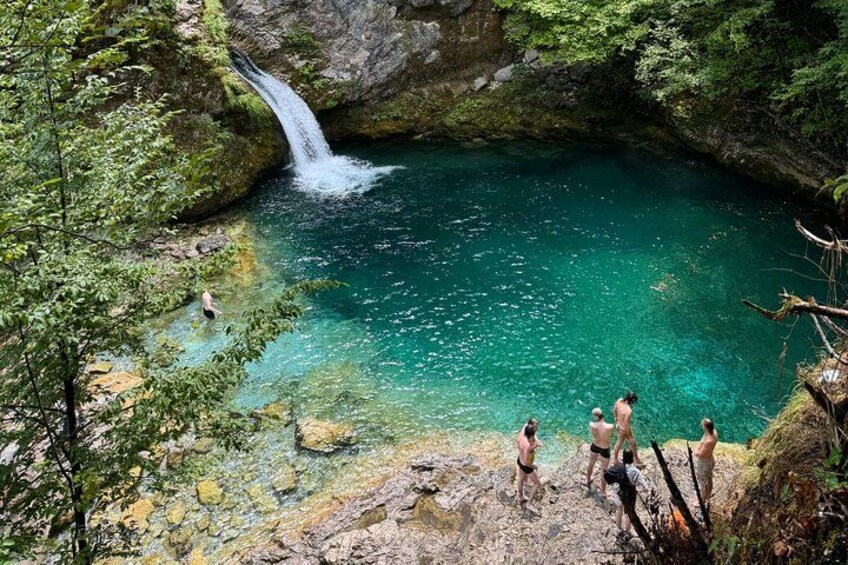 A waterfall, clear turquoise water, rocky terrain, and people standing on a cliff. 1