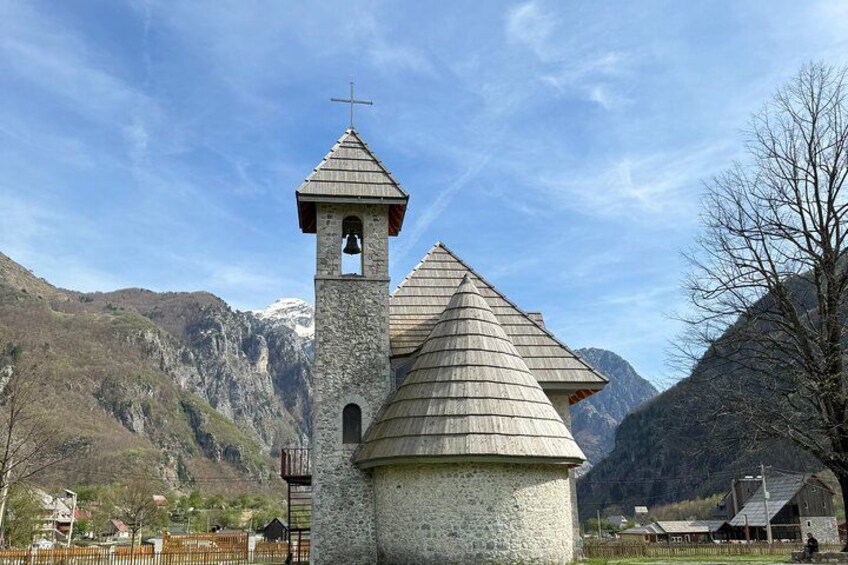 A church with a bell tower and a stone building with a conical roof, surrounded by mountains. 2