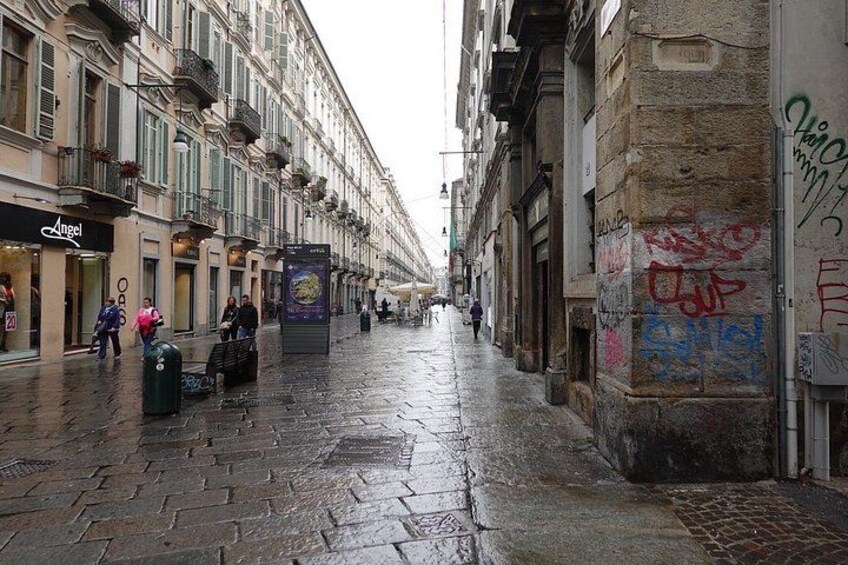 A wet cobblestone street lined with multi-story buildings, some with balconies and storefronts, including a visible 'Angel' shop. 4