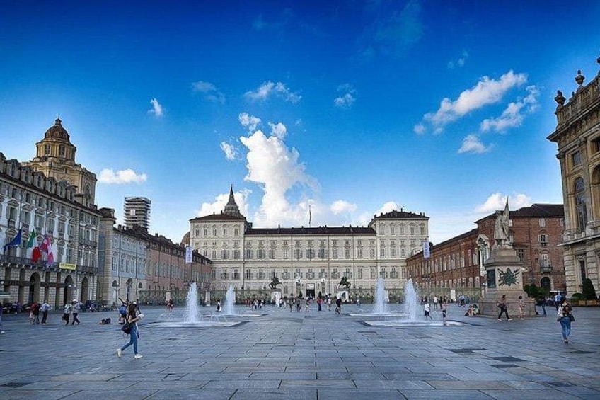A large open square with a historic building in the center, fountains, and people walking. 1