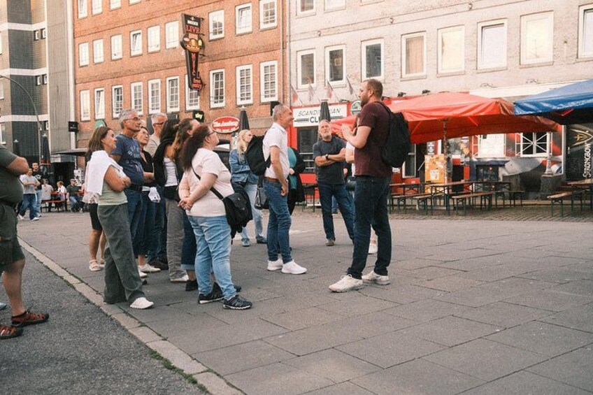 Een groep mensen staat op een stoep in een stedelijk gebied, met gebouwen en terrasjes op de achtergrond. 4