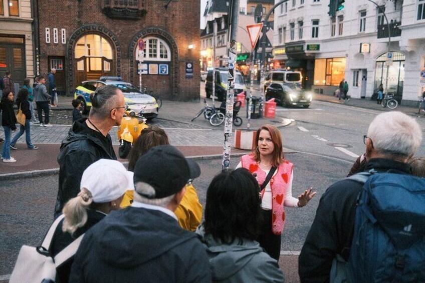 Een groep mensen staat op een hoek van een straat, met gebouwen en voertuigen op de achtergrond. 3