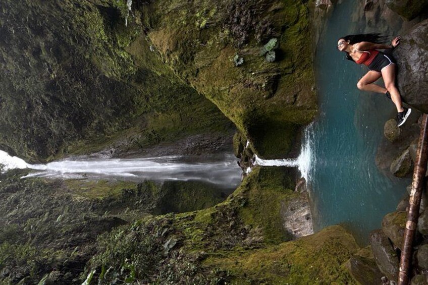 A waterfall cascading down a moss-covered cliff, a person standing on a rock ledge, and a clear blue pool at the base. 2
