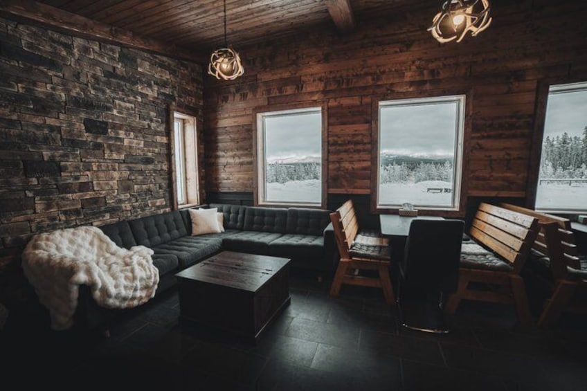 A rustic interior with a stone wall, wooden furniture, and large windows overlooking a snowy landscape. 1