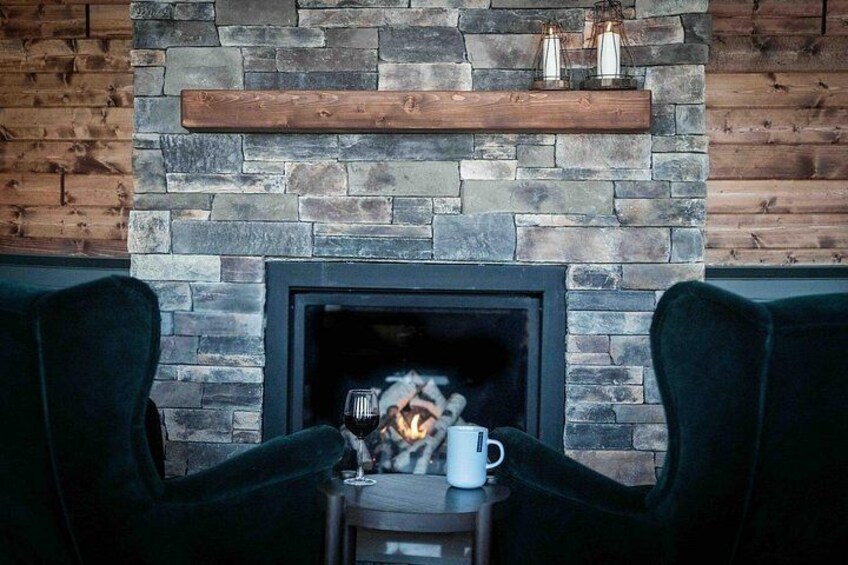 A fireplace with a stone wall, two chairs, a table with a mug, and a wooden shelf above the fireplace. 4