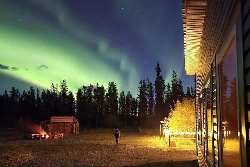 A person standing in a forested area at night with the Northern Lights in the sky, a building with illuminated windows, and a vehicle with its lights on. 3