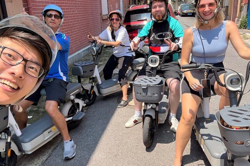 Fire personer på el-scootere, en person på en cykel og en rød bil parkeret på gaden. 1