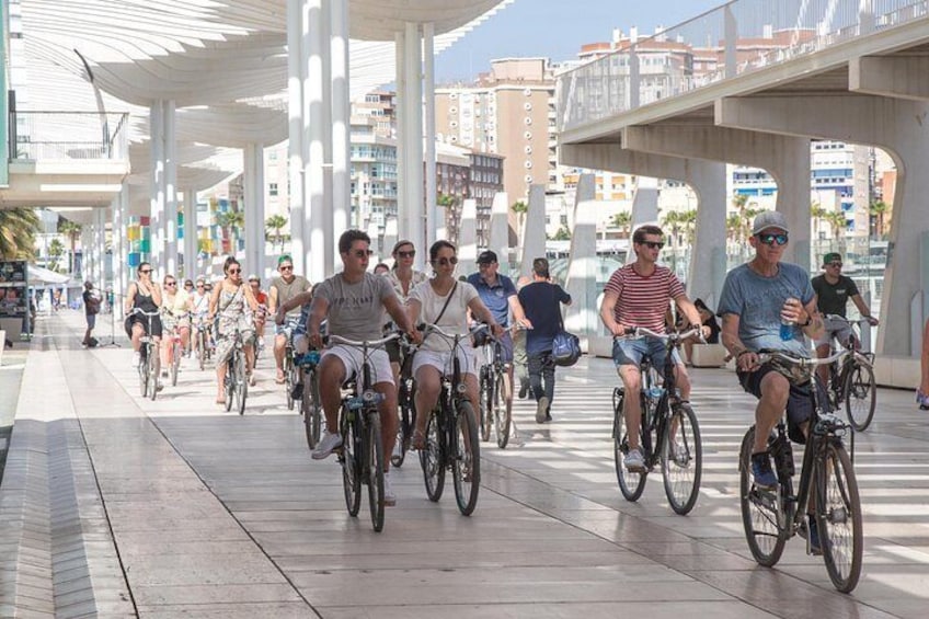 En gruppe cyklister cykler på en belagt sti under et overdækket gangareal med bygninger i baggrunden. 4