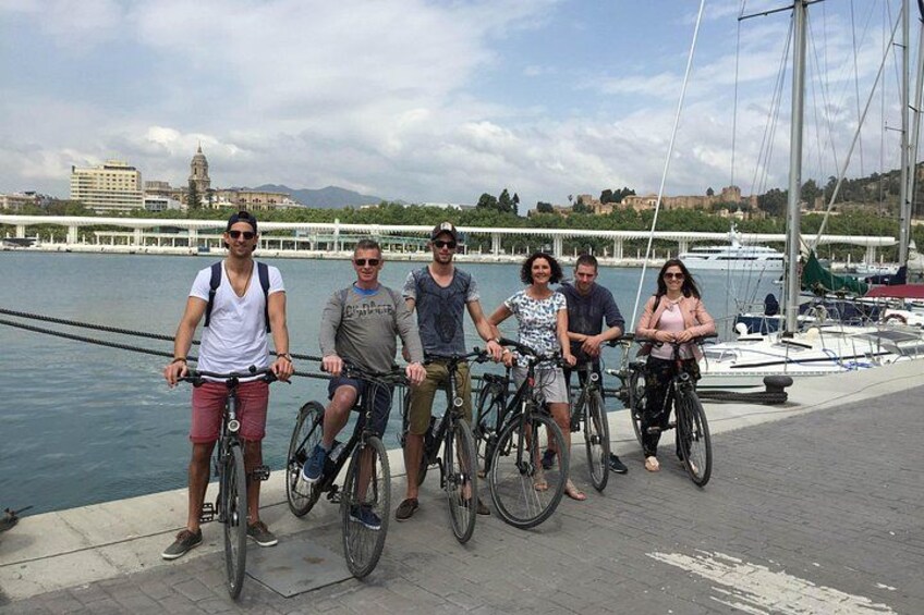 A group of people on bicycles by a waterfront with boats and a bridge in the background. 4