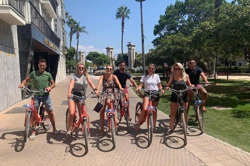 A group of people riding bicycles on a paved path with buildings and palm trees in the background. 2