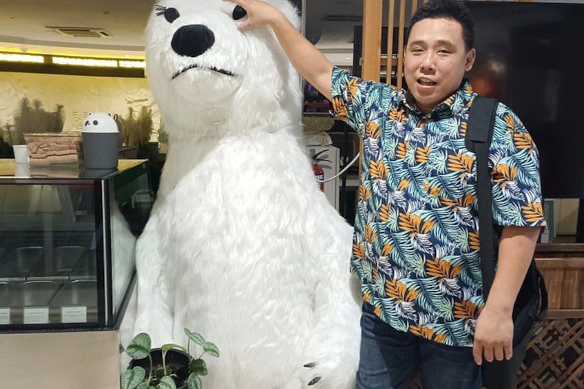 A person standing next to a large white bear statue in an indoor setting with a glass display case and a plant in the background. 3
