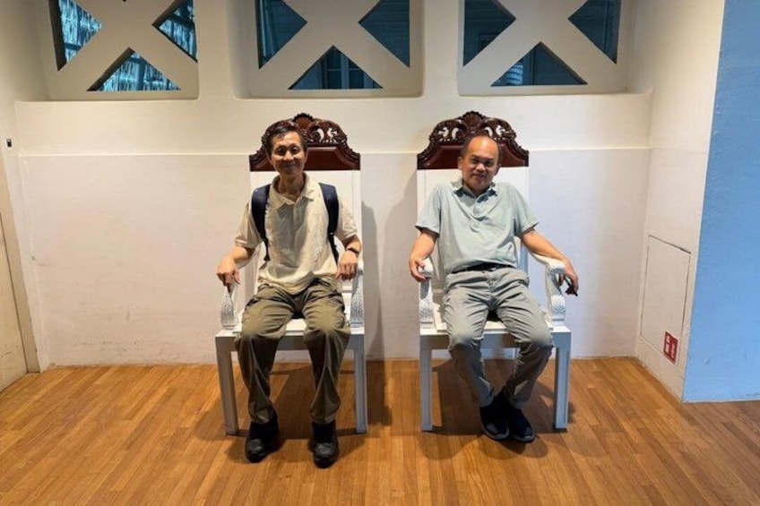 Two individuals seated on ornate chairs in a room with wooden flooring and a white wall with decorative window frames. 2