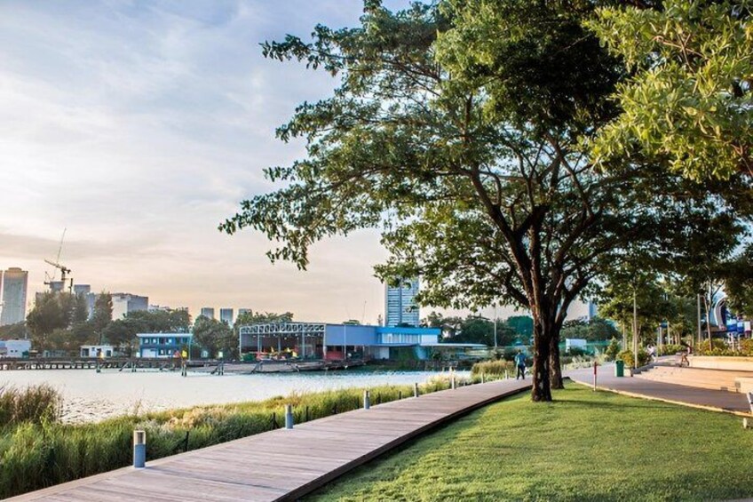 A riverside walkway with a tree, a grassy area, and buildings in the background. 1