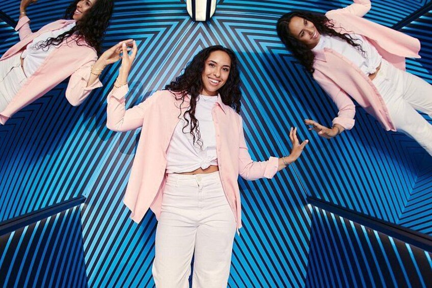 A person in a pink jacket and white pants standing in front of a blue geometric patterned background. 2