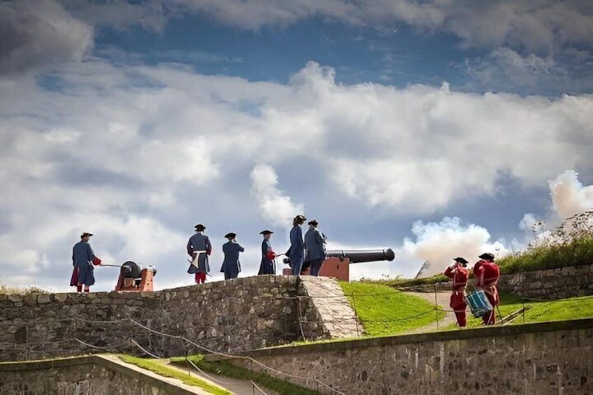 A historical reenactment with individuals in period costumes on a stone wall, a cannon, and a cloudy sky. 1